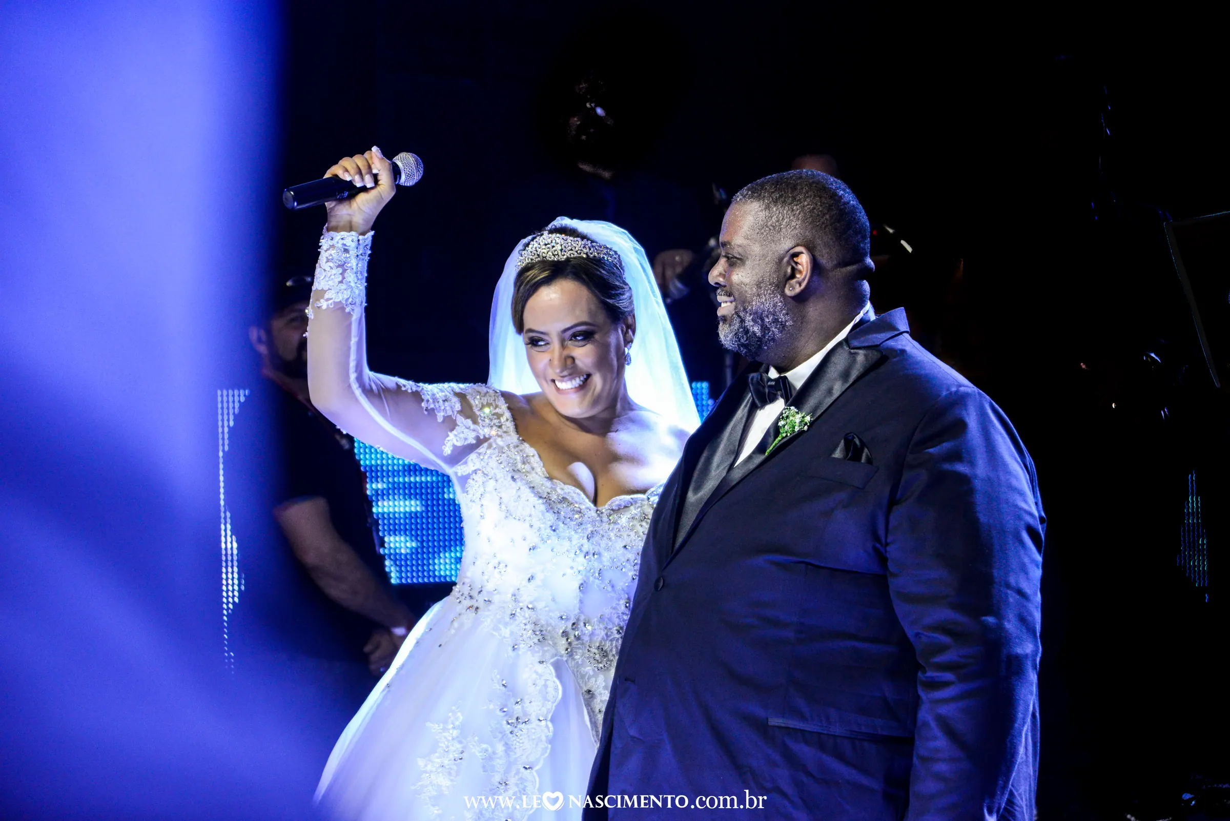 Casal de noivos celebrando no palco sob luz azul, com convidados ao fundo